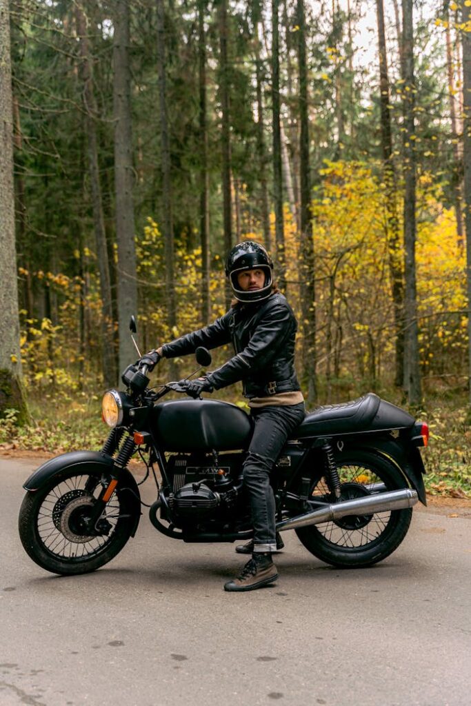 Motorcyclist wearing a black leather jacket and helmet sitting on a motorbike in a forest.